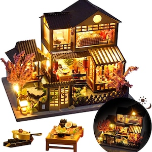 May include: Miniature Japanese-style wooden dollhouse with interior lighting. The two-story structure features a dark roof, detailed balconies, and miniature furniture. Includes a small table with food and a separate pot. Decorative cherry blossom branches add color.
