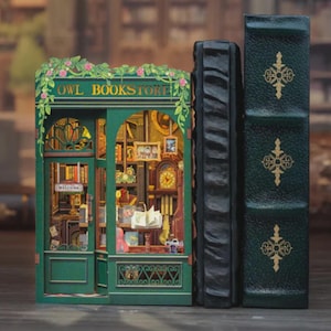 K&ouml;nnte beinhalten: Ein detailliertes, gr&uuml;nes Miniaturmodell der "Owl Bookstore" mit zwei Buchst&uuml;tzen. Die Buchhandlung hat eine Uhr, B&uuml;cher und den Text "WELCOME". Die Buchst&uuml;tzen sind schwarz und dunkelgr&uuml;n mit goldenen Akzenten. Das Modell ist ein Buchst&uuml;tzen-Set.