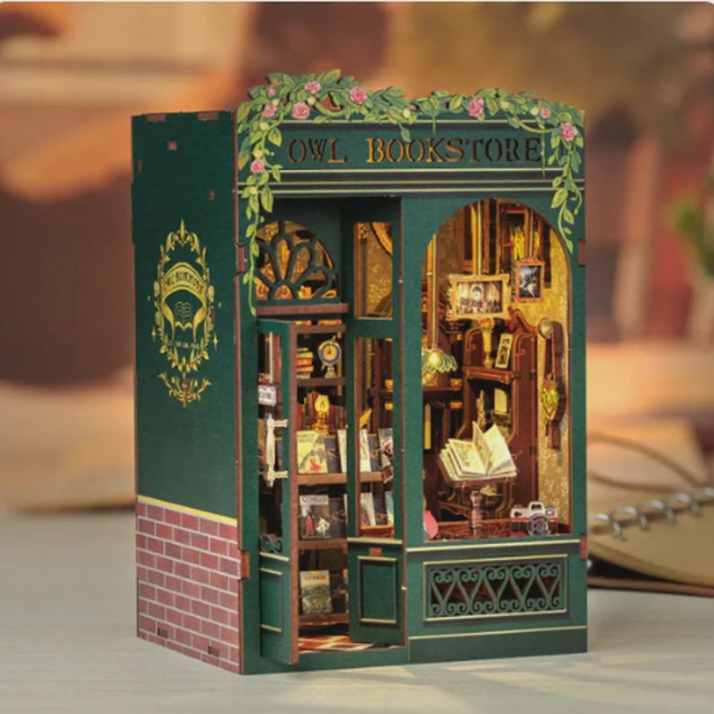 K&ouml;nnte beinhalten: Miniaturmodell der Owl Bookstore mit gr&uuml;ner Fassade, roter Ziegelbasis und floralen Akzenten. Der Laden verf&uuml;gt &uuml;ber detaillierte Innenelemente, darunter B&uuml;cherregale, B&uuml;cher und einen Lesebereich. Der Text "OWL BOOKSTORE" ist &uuml;ber dem Eingang zu sehen.