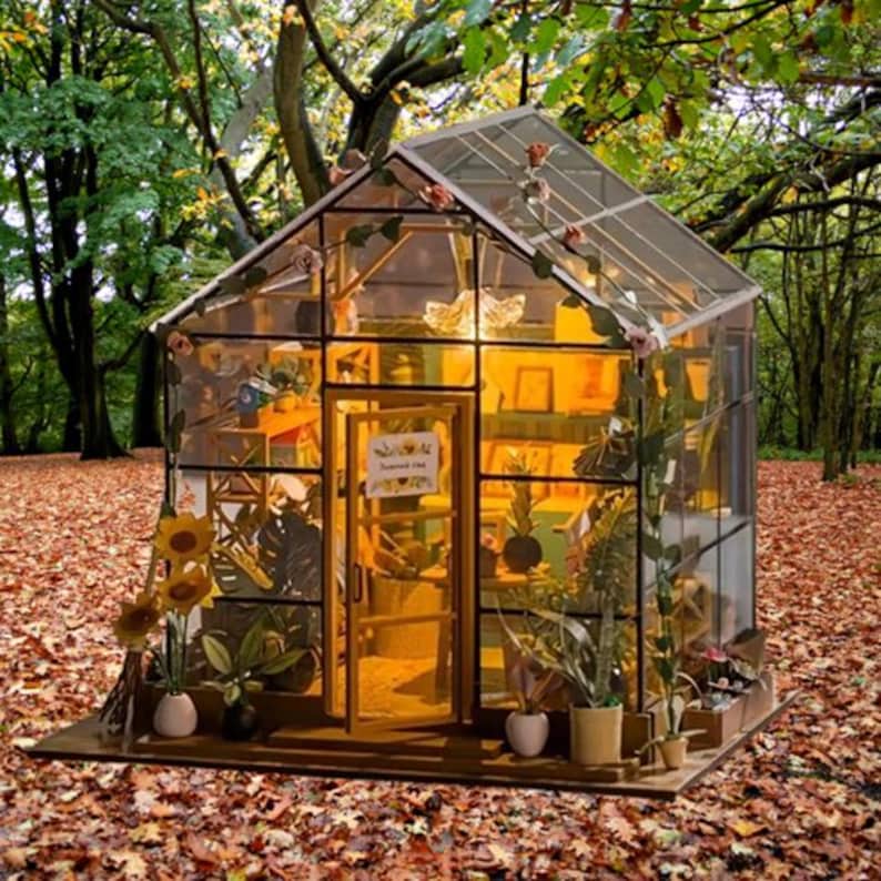 May include: A miniature greenhouse with a glass roof and walls, set amidst a backdrop of autumn leaves and trees. The structure features a wooden frame, a door, and is decorated with artificial flowers. Inside, miniature plants and decor are visible.