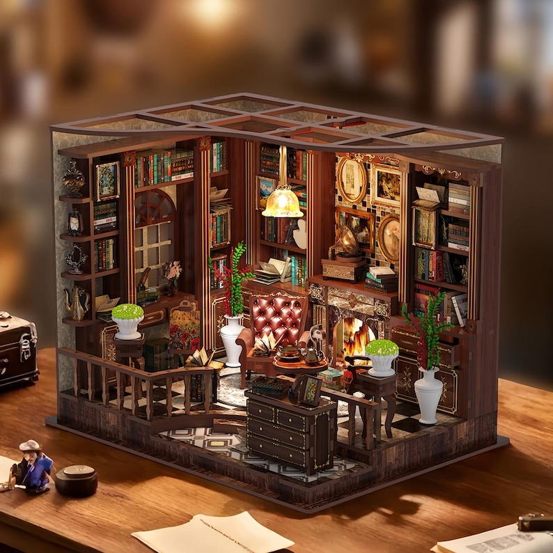 May include: A miniature wooden library diorama with detailed bookshelves, a fireplace, and a red armchair. The scene includes a small table, a lamp, and decorative items, all within a framed structure. The diorama is set on a wooden surface.