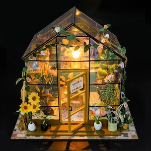 May include: Miniature greenhouse model with a glass roof and walls, featuring a yellow door and interior lighting. The structure is adorned with miniature plants, flowers, and vines. A small sign is visible on the door. The base is made of a patterned material.