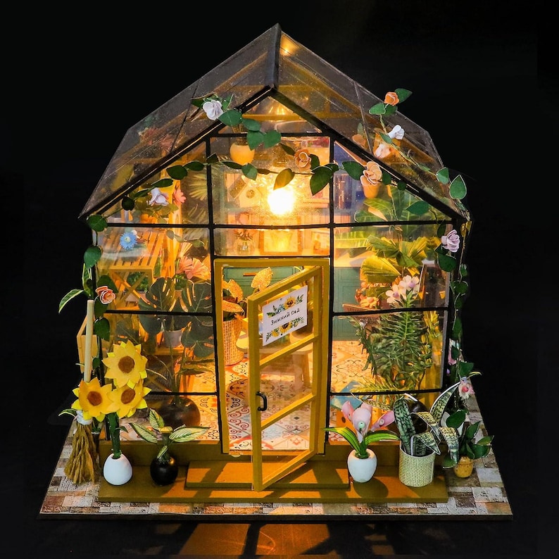 May include: Miniature greenhouse model with a glass roof and walls, featuring a yellow door and interior lighting. The structure is adorned with miniature plants, flowers, and vines. A small sign is visible on the door. The base is made of a patterned material.