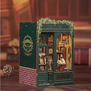 K&ouml;nnte beinhalten: Miniaturmodell der Owl Bookstore mit gr&uuml;ner Fassade, roter Ziegelbasis und aufwendigen Innendetails. Das Modell zeigt den Text "OWL BOOKSTORE" &uuml;ber dem Eingang, mit floralen Akzenten. B&uuml;cherregale, Uhren und Miniaturm&ouml;bel sind im Inneren sichtbar.