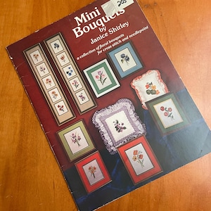 May include: A softcover instructional booklet titled Mini Bouquets by Janice Shirley, a collection of floral bouquets for cross stitch and needlepoint. The dark red cover features various framed floral embroidery projects and two small pillows.