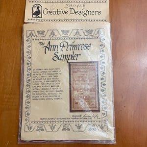 May include: A vintage cross-stitch sampler kit titled "The Ann Primrose Sampler" by Creative Designers. The kit includes imported linen, DMC floss, and illustrated instructions. The design features a framed sampler with text and decorative borders.