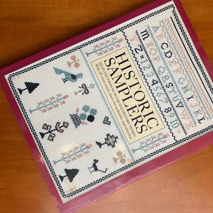 May include: A softcover book titled Historic Samplers with a magenta border. The cover features a white background with cross-stitch patterns including an alphabet, numbers, trees, birds, and flowers. The text reads Historic Samplers Selected From Museums And Historic Homes With 30 cross-stitch charts for authentic reproduction.