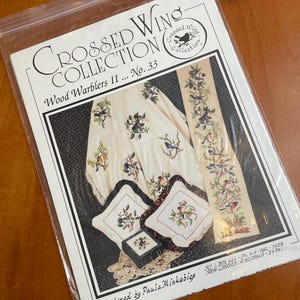 May include: A Crossed Wing Collection cross-stitch pattern booklet titled Wood Warblers II No. 33, designed by Paula Minkebige. The cover features images of embroidered fabric items including a cloth, two square pillows, and a long vertical runner.