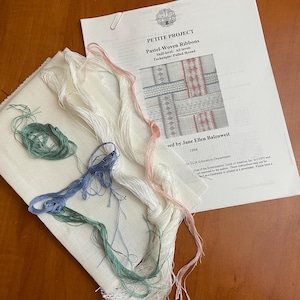 May include: A craft project with a white fabric square, colorful threads, and a printed instruction sheet. The threads are in shades of pink, blue, green, and white. The instruction sheet is titled "PETITE PROJECT Pastel-Woven Ribbons."