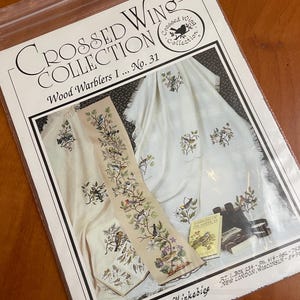 May include: A clear plastic bag containing a cross-stitch pattern titled Crossed Wing Collection Wood Warblers I No. 31. The cover features images of embroidered fabric with bird designs, a book titled Warblers of the Americas, and text.