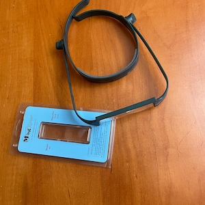 May include: A dark green head-mounted magnifier with a circular frame and a rectangular lens. The product is packaged in a blue and clear plastic sleeve with the brand name "MagEyes" printed on it.