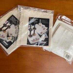 May include: Three packages of Country Cottage Table Linens, featuring a floral embroidery design. The linens are packaged in clear plastic, with a cream-colored fabric and a fringed edge. The packaging includes the brand name "Janlynn".