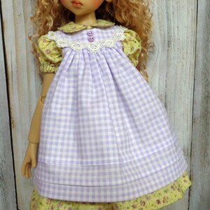 May include: A doll wearing a purple and white gingham dress with a yellow floral print underneath. The doll has blonde curly hair and is wearing a green and purple striped bow. The doll is wearing white socks with lace trim and purple shoes with lace trim.