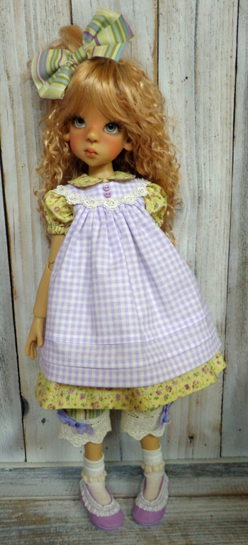 May include: A doll wearing a purple and white gingham dress with a yellow floral print underneath. The doll has blonde curly hair and is wearing a green and purple striped bow. The doll is wearing white socks with lace trim and purple shoes with lace trim.
