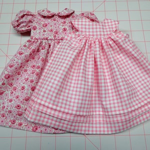 May include: Two pink doll dresses. One dress has a floral print and the other has a gingham pattern.