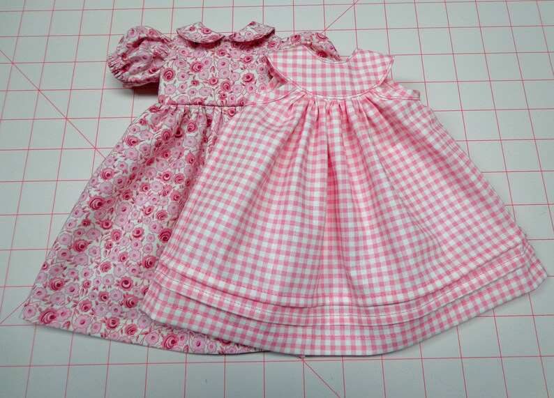 May include: Two pink doll dresses. One dress has a floral print and the other has a gingham pattern.