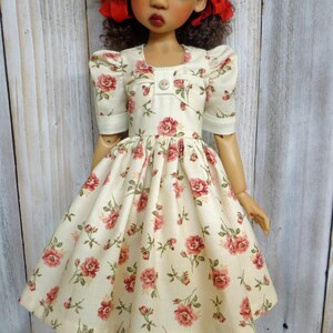 May include: A doll wearing a white dress with a floral pattern and red bows in her hair. She is wearing white socks and light blue shoes.