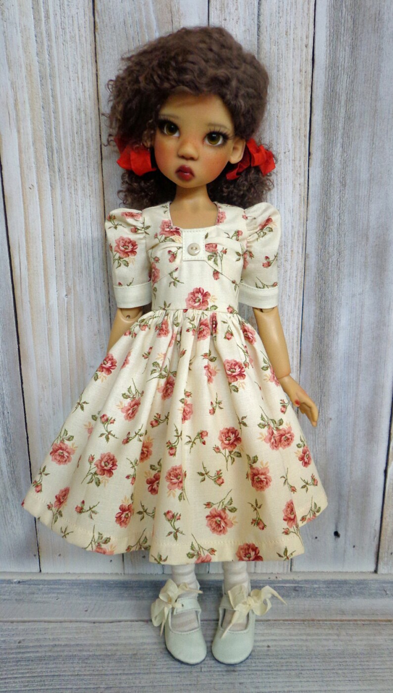 May include: A doll wearing a white dress with a floral pattern and red bows in her hair. She is wearing white socks and light blue shoes.
