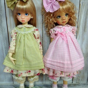 May include: Two dolls wearing gingham dresses with floral accents. The doll on the left is wearing a green dress with a green bow in her hair. The doll on the right is wearing a pink dress with a pink and white striped bow in her hair.