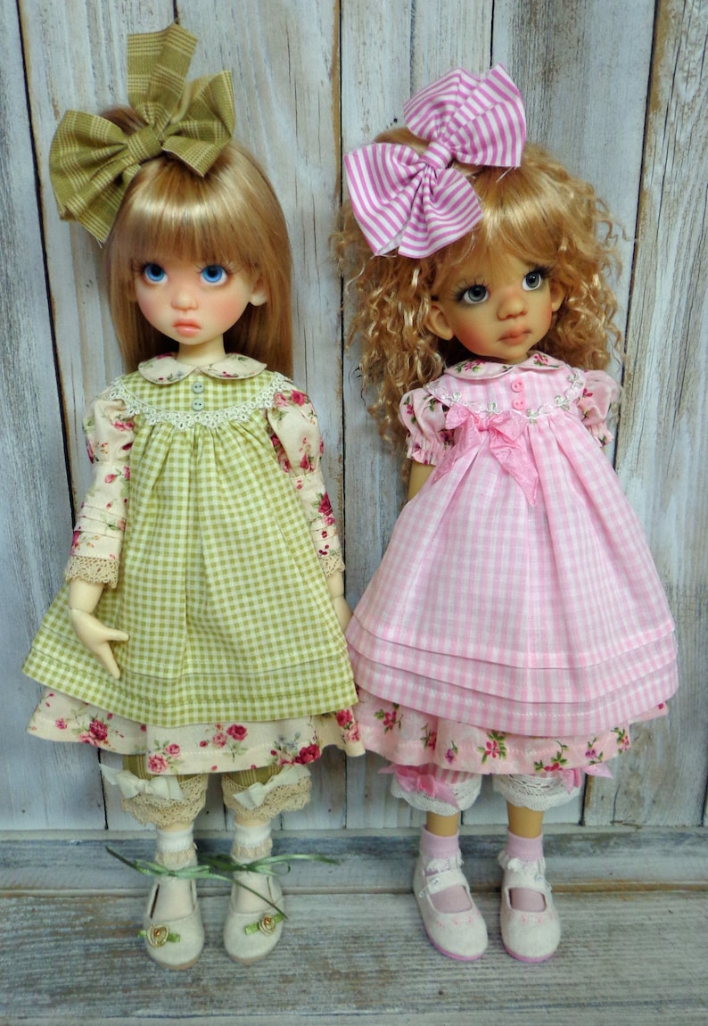 May include: Two dolls wearing gingham dresses with floral accents. The doll on the left is wearing a green dress with a green bow in her hair. The doll on the right is wearing a pink dress with a pink and white striped bow in her hair.