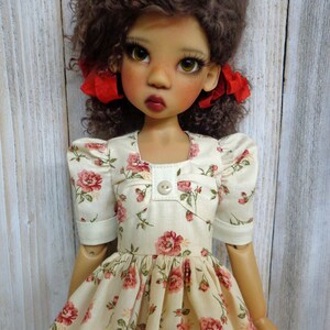 May include: A doll with brown curly hair wearing a cream-colored dress with a floral pattern and red bows in her hair.