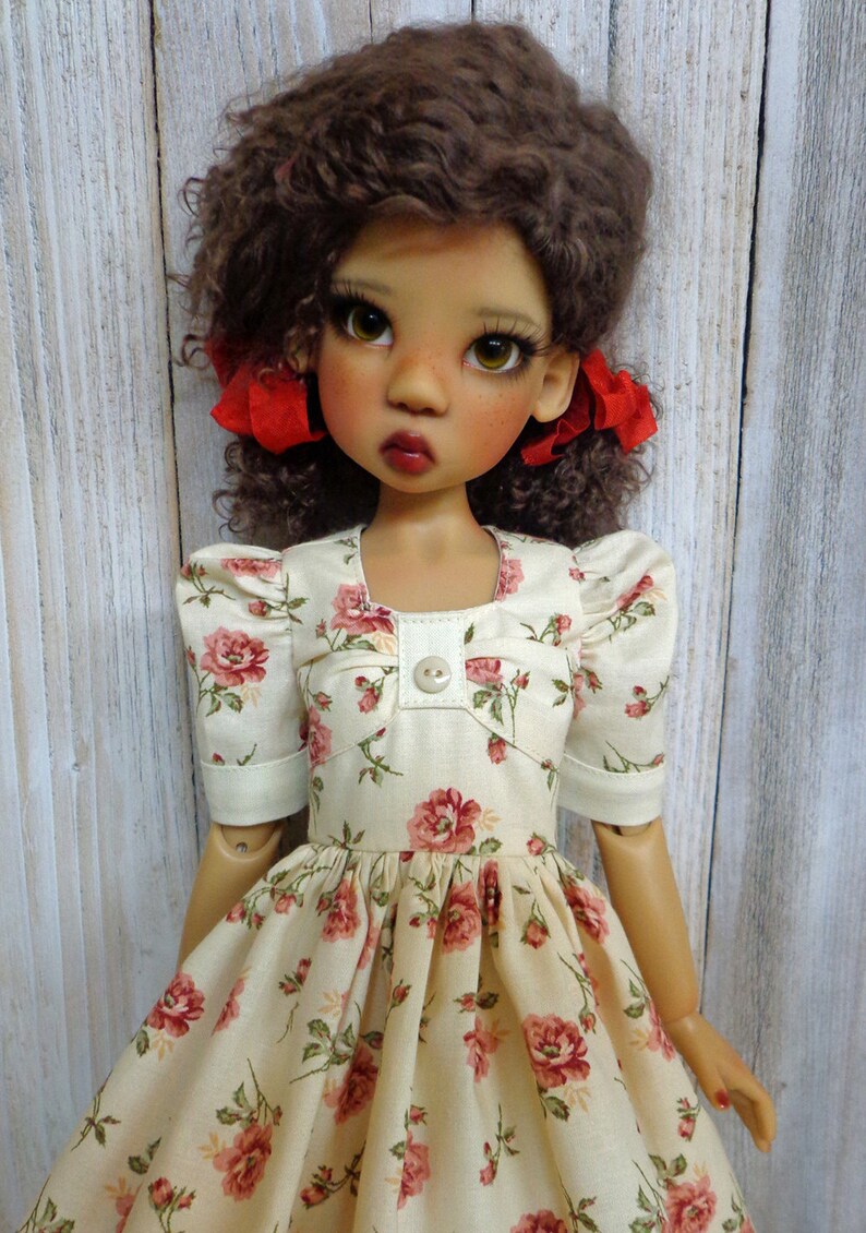 May include: A doll with brown curly hair wearing a cream-colored dress with a floral pattern and red bows in her hair.