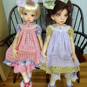May include: Two dolls wearing gingham dresses with floral trim and matching bows. The doll on the left is wearing a pink and white gingham dress and the doll on the right is wearing a purple and white gingham dress.