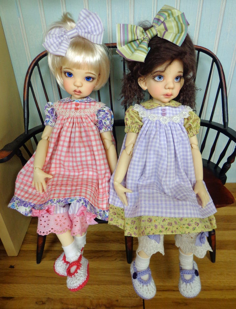 May include: Two dolls wearing gingham dresses with floral trim and matching bows. The doll on the left is wearing a pink and white gingham dress and the doll on the right is wearing a purple and white gingham dress.