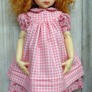 May include: A doll with red curly hair wearing a pink and white gingham dress with a pink bow in her hair. The doll is wearing pink socks and pink shoes with floral embroidery.