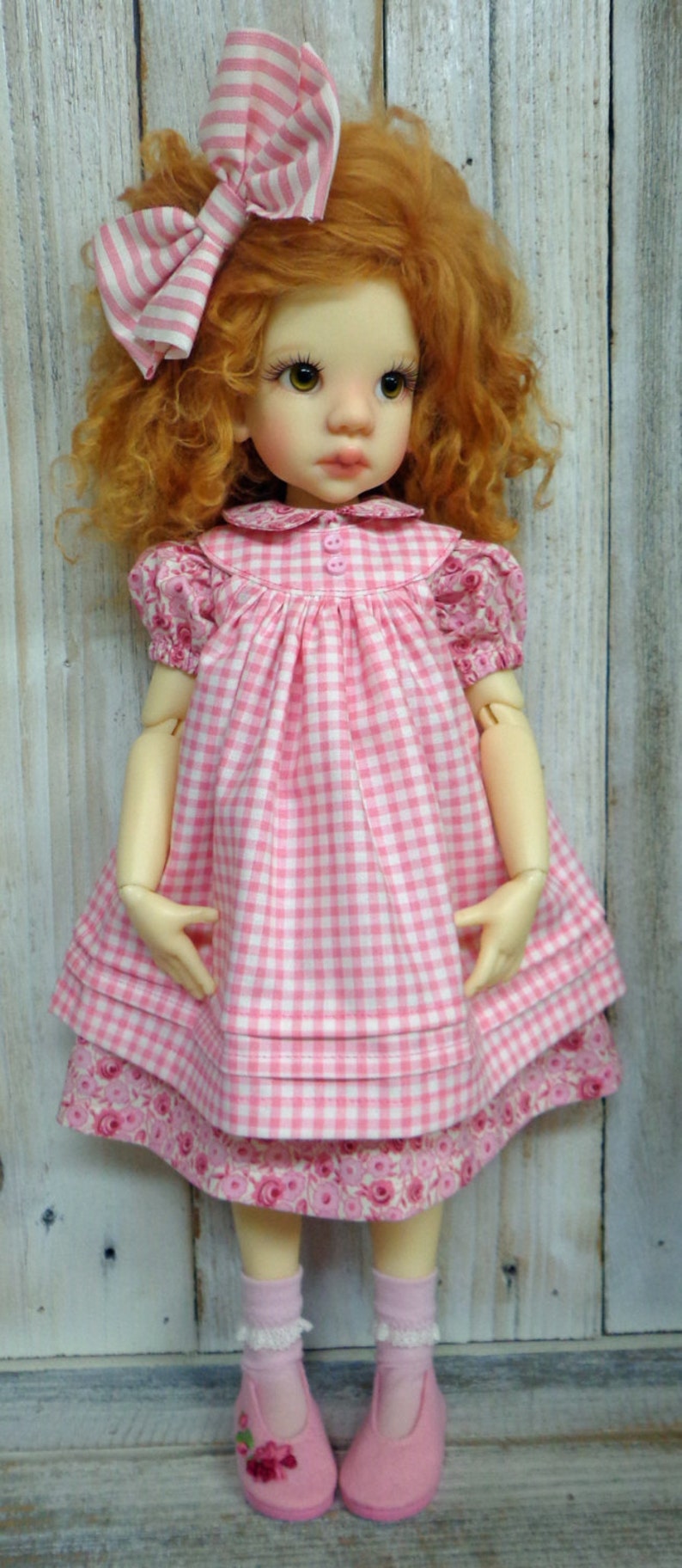 May include: A doll with red curly hair wearing a pink and white gingham dress with a pink bow in her hair. The doll is wearing pink socks and pink shoes with floral embroidery.