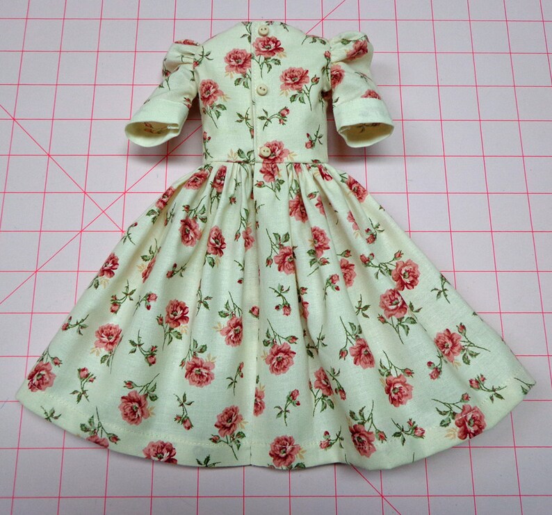 May include: A light green dress with a floral pattern of pink roses. The dress has a gathered skirt and puffed sleeves. It has a button closure down the back.