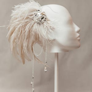 Elegant handmade feather headdress inspired by the Great Gatsby era, featuring soft white ostrich and dramatic rooster feathers. Embellished with pearls, lace, and sparkling crystals, this bridal headpiece adds a vintage, boho-chic touch to any wedding, festival, or photoshoot. Displayed on a white mannequin, the piece showcases fine craftsmanship, ideal for brides, performers, or themed events. Perfect as a wedding fascinator or statement hair accessory.