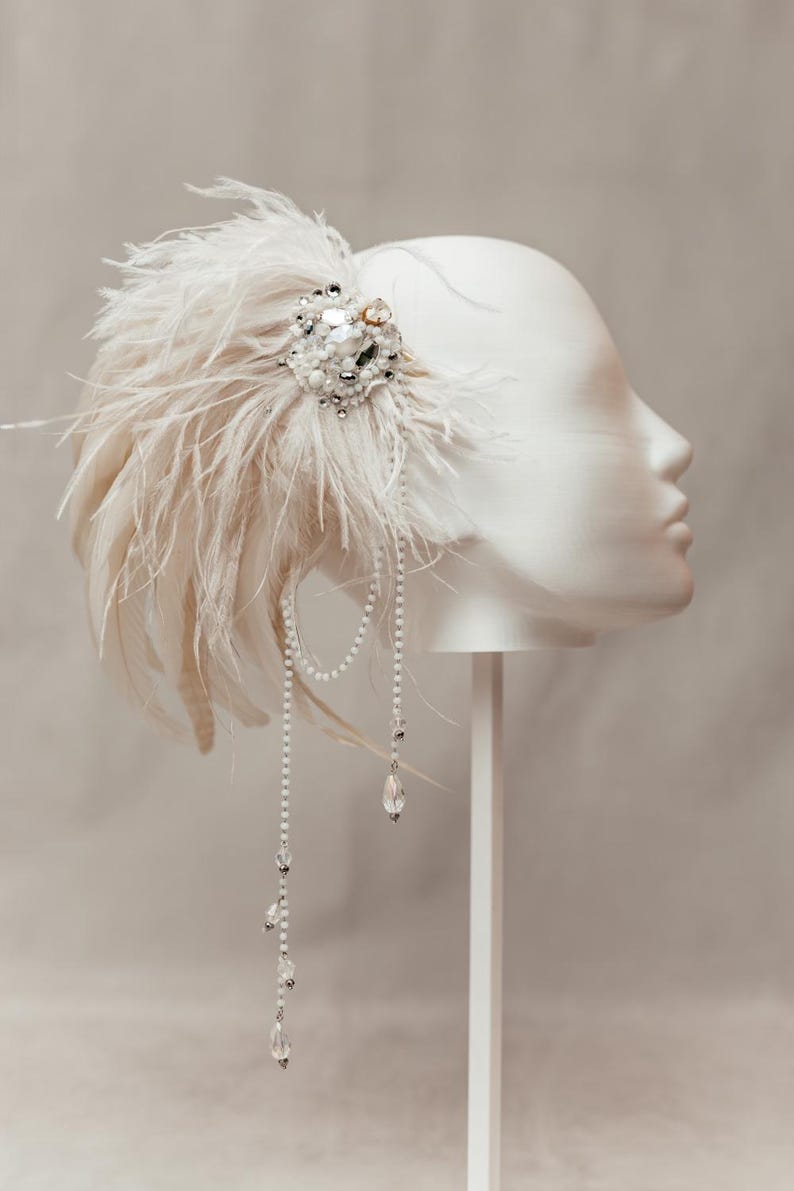 Elegant handmade feather headdress inspired by the Great Gatsby era, featuring soft white ostrich and dramatic rooster feathers. Embellished with pearls, lace, and sparkling crystals, this bridal headpiece adds a vintage, boho-chic touch to any wedding, festival, or photoshoot. Displayed on a white mannequin, the piece showcases fine craftsmanship, ideal for brides, performers, or themed events. Perfect as a wedding fascinator or statement hair accessory.