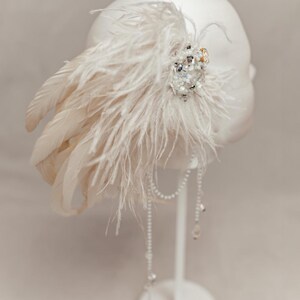 Elegant handmade feather headdress inspired by the Great Gatsby era, featuring soft white ostrich and dramatic rooster feathers. Embellished with pearls, lace, and sparkling crystals, this bridal headpiece adds a vintage, boho-chic touch to any wedding, festival, or photoshoot. Displayed on a white mannequin, the piece showcases fine craftsmanship, ideal for brides, performers, or themed events. Perfect as a wedding fascinator or statement hair accessory.