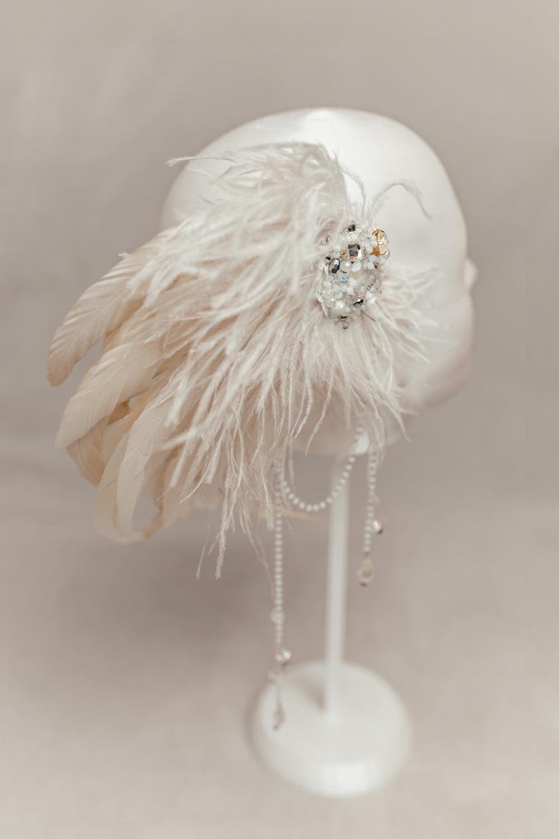 Elegant handmade feather headdress inspired by the Great Gatsby era, featuring soft white ostrich and dramatic rooster feathers. Embellished with pearls, lace, and sparkling crystals, this bridal headpiece adds a vintage, boho-chic touch to any wedding, festival, or photoshoot. Displayed on a white mannequin, the piece showcases fine craftsmanship, ideal for brides, performers, or themed events. Perfect as a wedding fascinator or statement hair accessory.