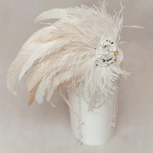 Dramatic feather headdress designed for carnival, fantasy costumes, or stage performance, handcrafted with lush white ostrich feathers and dark rooster accents. Embellished with shimmering pearls, crystals, and lace detailing, this statement hairpiece captures a bold, ethereal aesthetic. Ideal for masquerade balls, themed photo shoots, or artistic expression. Shown on a mannequin with neutral background to highlight the texture, movement, and layered design of the headpiece.