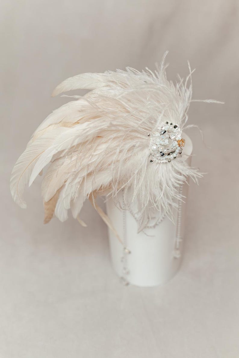 Dramatic feather headdress designed for carnival, fantasy costumes, or stage performance, handcrafted with lush white ostrich feathers and dark rooster accents. Embellished with shimmering pearls, crystals, and lace detailing, this statement hairpiece captures a bold, ethereal aesthetic. Ideal for masquerade balls, themed photo shoots, or artistic expression. Shown on a mannequin with neutral background to highlight the texture, movement, and layered design of the headpiece.