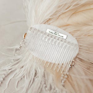 Elegant handmade feather headdress inspired by the Great Gatsby era, featuring soft white ostrich and dramatic rooster feathers. Embellished with pearls, lace, and sparkling crystals, this bridal headpiece adds a vintage, boho-chic touch to any wedding, festival, or photoshoot. Displayed on a white mannequin, the piece showcases fine craftsmanship, ideal for brides, performers, or themed events. Perfect as a wedding fascinator or statement hair accessory.