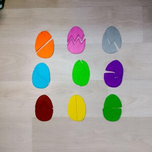 May include: Nine colorful felt Easter eggs, each cut into two pieces, arranged in three rows. The eggs are in various colors: orange, pink, gray, blue, green, purple, maroon, yellow, and green. The pink egg has a zig-zag design.