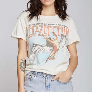 May include: An off-white short-sleeve t-shirt with raw-edge sleeves and neckline. The front features a graphic of a winged figure in flight with the text "LED ZEPPELIN" in orange and "UNITED STATES OF AMERICA TOUR 1977" in black lettering.
