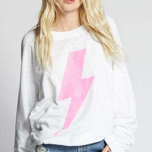 May include: White long-sleeve shirt featuring a large pink lightning bolt graphic. The shirt is worn with denim shorts. The overall style is casual and youthful, suitable for everyday wear.