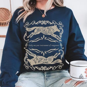 Op de afbeelding: Marineblauwe sweatshirt met een decoratief ontwerp met een wolf en een haas, omlijst door sierlijke krullen. De tekst "Make me your instrument of vengeance" staat gecentreerd onder de wolf. De sweatshirt is effen van kleur.