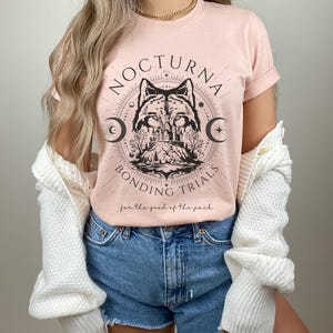May include: A light pink t-shirt featuring a circular graphic with a wolf illustration, crescent moons, and the words "NOCTURNA BONDING TRIALS" in black. The phrase "for the good of the pack" is printed below the graphic. The shirt has short sleeves.