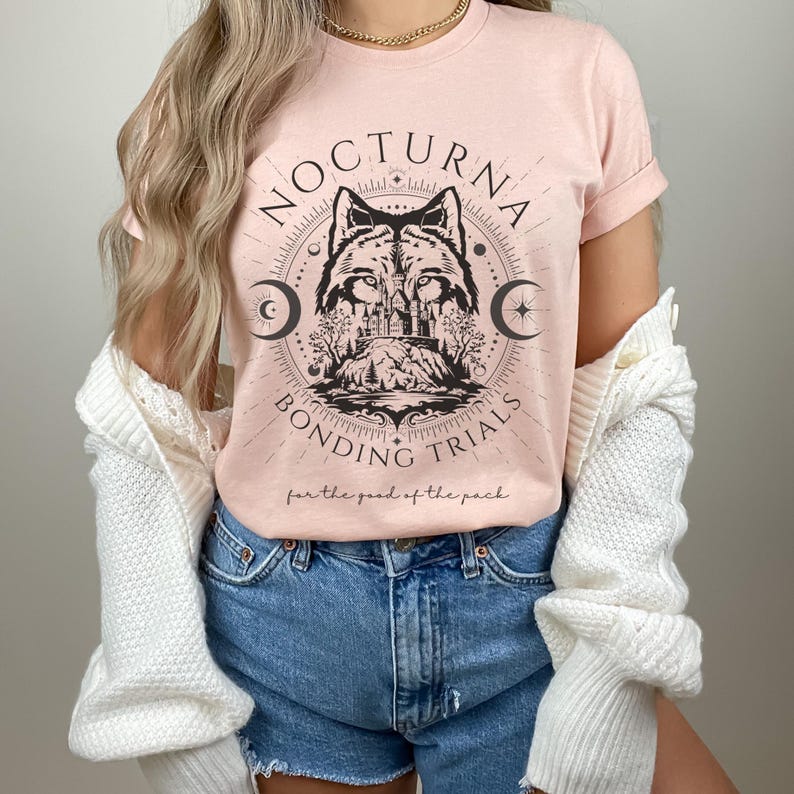 May include: A light pink t-shirt featuring a circular graphic with a wolf illustration, crescent moons, and the words "NOCTURNA BONDING TRIALS" in black. The phrase "for the good of the pack" is printed below the graphic. The shirt has short sleeves.