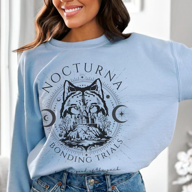 May include: Light blue sweatshirt with a circular graphic featuring a wolf, castle, and text that reads "NOCTURNA BONDING TRIALS". The design includes crescent moons and a mountain landscape.