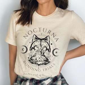 May include: Cream-colored t-shirt featuring a circular graphic with a wolf's head, a castle, and the words "NOCTURNA BONDING TRIALS" and "for the good of the pack." The design is in black ink.