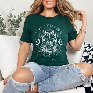 May include: A dark green t-shirt with a white graphic featuring a wolf, crescent moons, and the text "NOCTURNA BONDING TRIALS". The shirt also includes the text "for the good of the pack". The person is wearing ripped blue jeans.