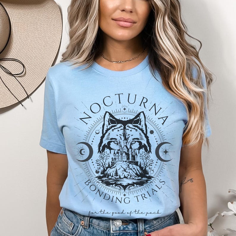 May include: Light blue t-shirt with a circular graphic featuring a wolf, castle, and crescent moons. The text "NOCTURNA BONDING TRIALS" and "for the good of the pack" are also displayed. The shirt is a crew neck.