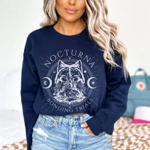 May include: Navy blue sweatshirt with a circular graphic featuring a wolf, castle, and crescent moons. The text "NOCTURNA BONDING TRIALS" is printed around the design in a light gray color. The sweatshirt is a crew neck style.