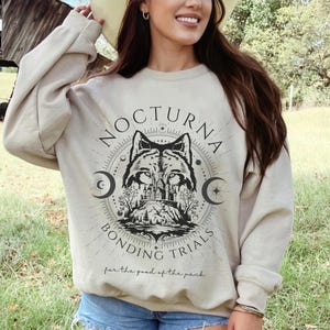 May include: Beige crewneck sweatshirt with a black and white wolf graphic. The graphic includes the text "NOCTURNA BONDING TRIALS" and "for the good of the pack". The design features a wolf, trees, and crescent moons.